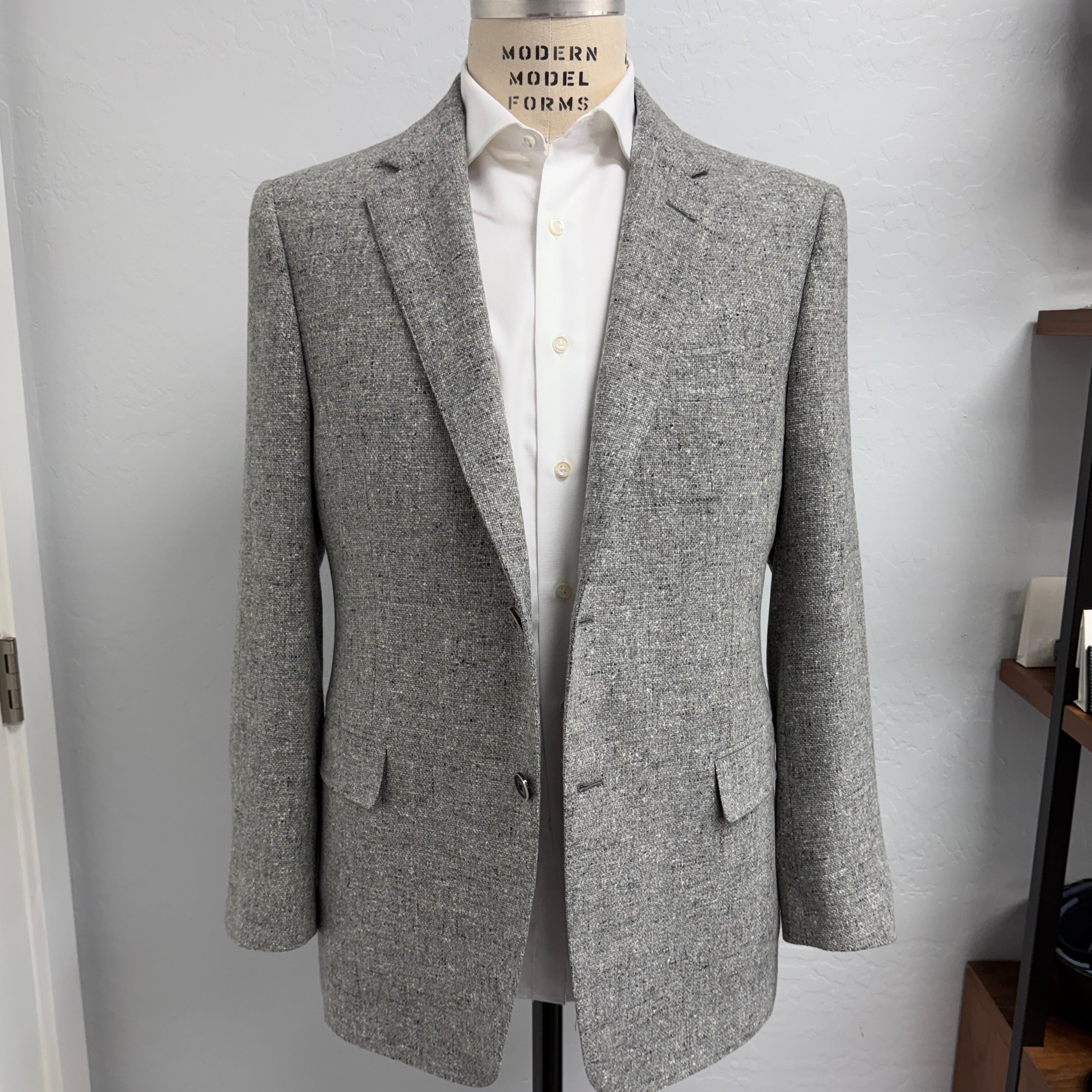 A light gray Loro Piana wool blazer displayed on a dress form mannequin, layered over a white button-down shirt. The blazer features notch lapels, a single-breasted button closure, and flap pockets, set against a plain light gray wall with a wooden shelf visible on the right.
