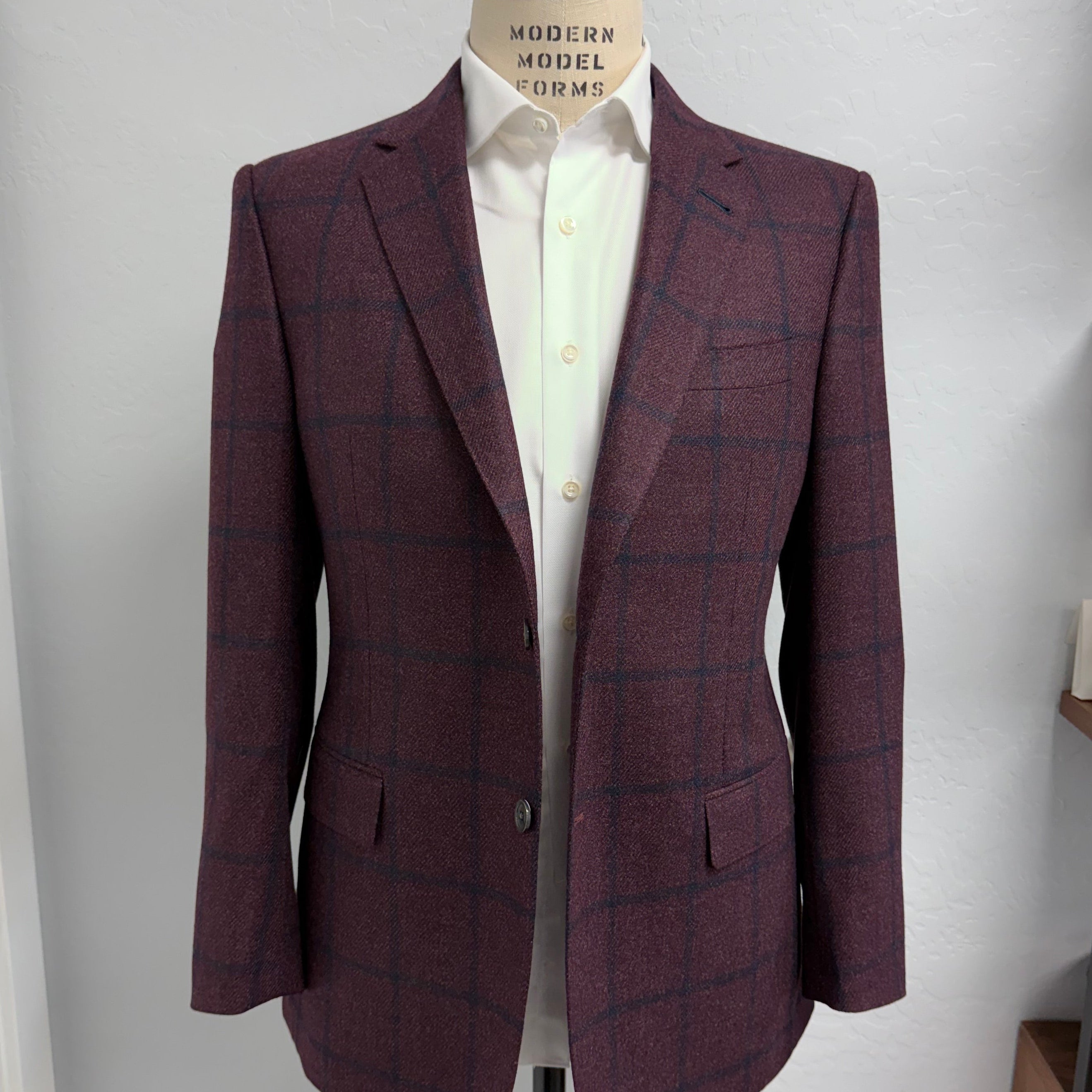 A Loro Piana men's single-breasted blazer in a burgundy and navy windowpane check tweed, displayed on a dress form mannequin wearing a white dress shirt. The blazer is shown against a plain white wall with a wooden shelf visible on the right.