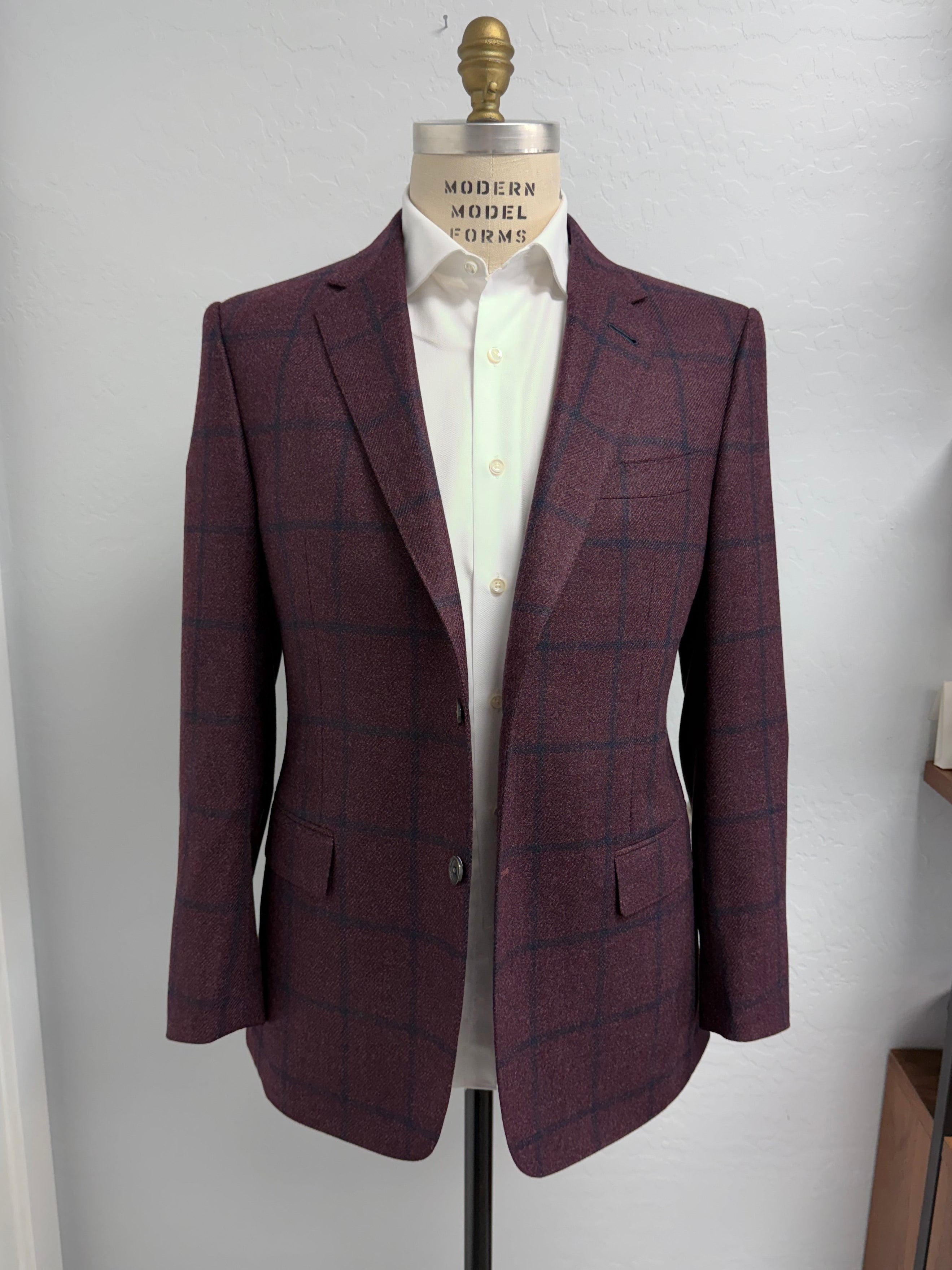 A Loro Piana men's single-breasted blazer in a burgundy and navy windowpane check tweed, displayed on a dress form mannequin wearing a white dress shirt. The blazer is shown against a plain white wall with a wooden shelf visible on the right.